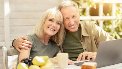 A smiling couple sits closely at a table outdoors, working on a laptop. Fresh fruits and snacks surround them as they enjoy the warm sunlight in a cozy setting, creating a joyful atmosphere.