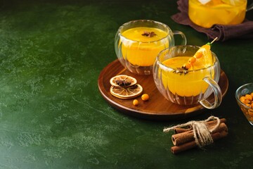 Sea buckthorn tea in clear cups with sea buckthorn berries and orange slices on a brown tray against a green concrete background.