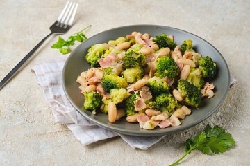 Fried broccoli with bacon and canned white beans on a gray plate against a light concrete background. Broccoli recipes.