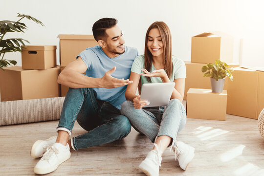 A couple sits on the floor surrounded by moving boxes, smiling and engaged in conversation. They hold a tablet while planning their next steps in organizing their new space.