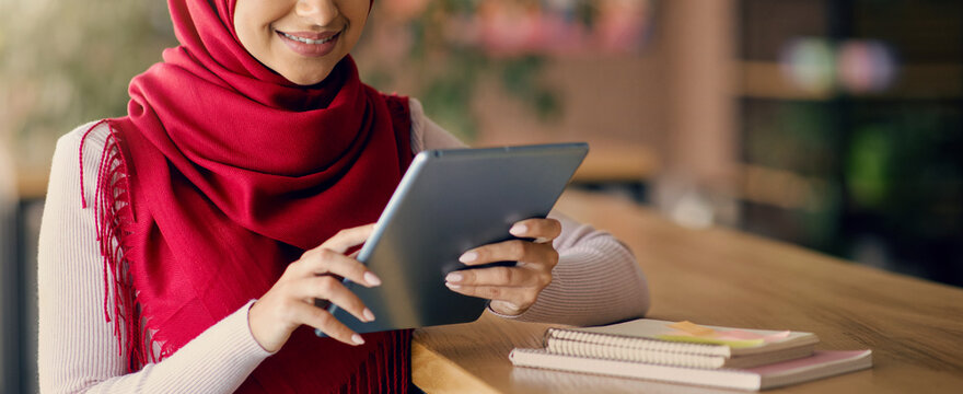 A woman wearing a red scarf is focused on her tablet while seated at a wooden table in a cozy café. Notebooks are placed beside her, suggesting she is studying or working. - Powered by Adobe