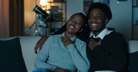 A young couple sits close on the couch, delighted by a romantic movie. The guy embraces his girlfriend as they chat, laugh, and share a cozy and joyful moment.
