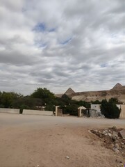 A sandy pathway leading toward the pyramids of Giza, framed by scattered trees and a cloudy sky, capturing the raw and authentic atmosphere of the ancient desert surroundings.