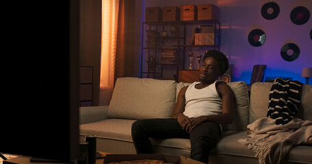 Obraz premium A pensive man of different nationalities dressed in a white T-shirt with straps sits on a sofa, quietly staring into the distance, showing signs of sadness and personal reflection.