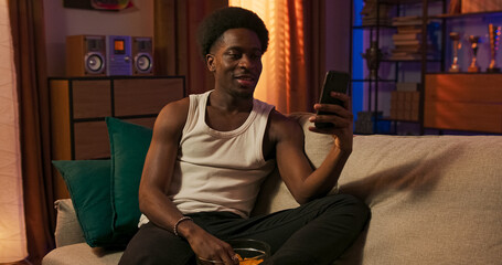 A casual man with a muscular build relaxes on the couch, messaging on his phone with a calm face, illuminated by modern LED lights, chips nearby on the table.