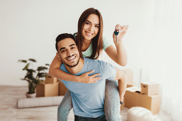 A happy couple stands in their new home, with one partner giving a piggyback ride. They are smiling while holding a set of keys, surrounded by moving boxes and light decor.