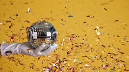 Gilded confetti falling on a hand in a white glove holding a mirror disco ball against a vibrant yellow background, creating a festive and glamorous party or celebration atmosphere - Powered by Adobe