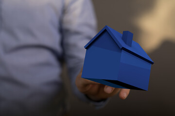 A person's hand holding a blue model house, representing home ownership, real estate, and housing...