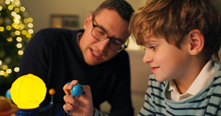 Close-up of seven-year-old boy and dad, who are considering model of the solar system. The boy is holding model of the planet Earth, and father with glasses tells him about it. Boy listens intently.