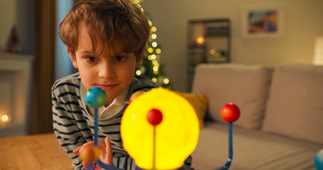 A smiling handsome boy looks at a children's model of the solar system. He studies the planets and smiles, wearing a shirt and a striped sweater. A Christmas tree flickers behind.