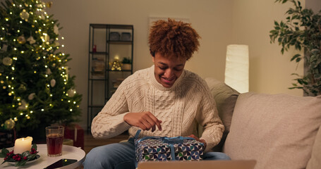 A boy in a sweater sits on a couch in a cozy apartment with a Christmas tree and decorations. He talks with friends through the camera on a laptop and unwraps a live gift he received from them.