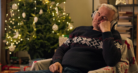 After Christmas party, exhausted middle-aged man sat down in chair by the Christmas tree.The man listens to the audiobook in headphones, rests head on hand and closes eyes. He is wearing warm sweater.