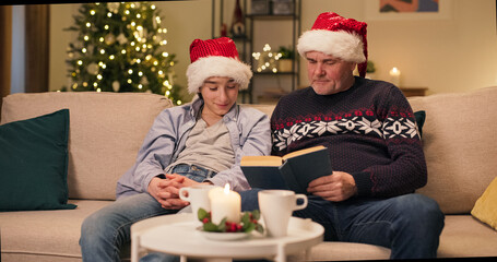 On the eve of Christmas, a teenager sits on a couch with his dad, who reads him a favorite book. The young man listens intently. They are wearing Santa hats, smiling and looking at the camera.
