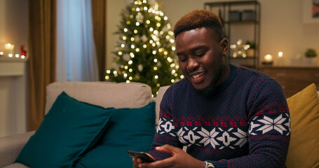 A young cheerful guy sits on the couch and talks on the phone. He communicates with girlfriend and...