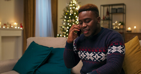 A young man of multi-racial background sits on a couch on a winter evening, emotionally engaged on the phone. He gestures with his hands and looks nervous, dressed in a warm sweater and a ball earring