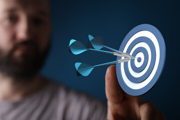 A close-up shot of a man's hand holding a dartboard with three darts hitting the bullseye,...
