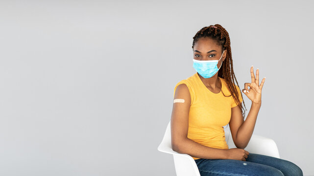 A young black woman in a yellow shirt and face mask gives an okay gesture after being vaccinated against the coronavirus. She poses confidently in a gray studio background.