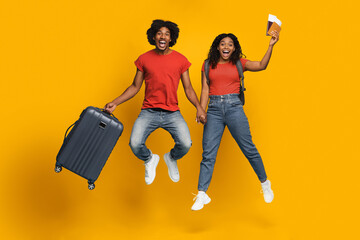 A happy couple is jumping against a bright yellow background, holding hands. One partner carries a...