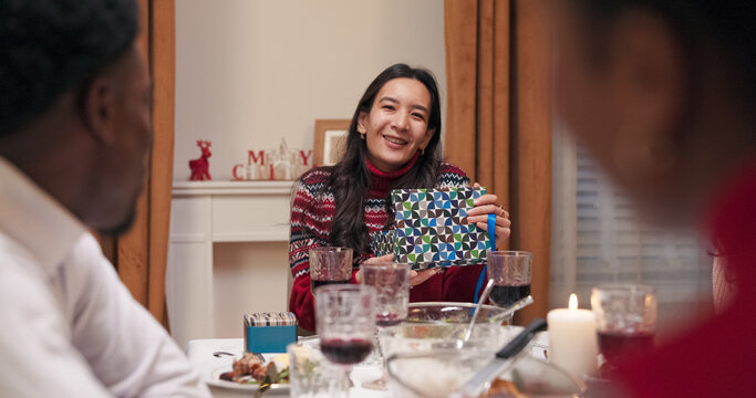 Friends laugh, chat, and sit at the festive table, exchanging gifts. A girl with long black hair was given a Christmas present; she was happy, untied the blue ribbon, opened the gift, and smiled.