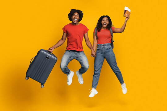 A happy couple is jumping against a bright yellow background, holding hands. One partner carries a suitcase while the other holds travel papers, suggesting excitement for their journey.
