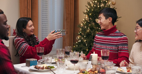Wearing the same sweaters, a girl with long black hair hands a gift to a boy at the festive table. The man opens the gift, rejoices, and smiles at the camera with his friends.