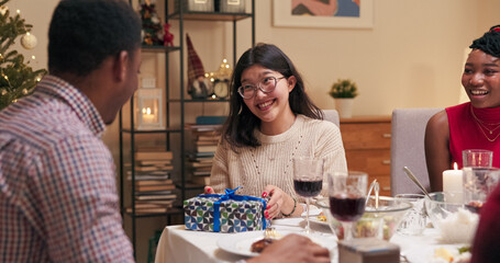 Friends sit at the festive table and take turns giving Christmas gifts. Girl with black hair and glasses gives gift to guy in shirt. The student happily accepts the gift, opens the box and smiles.