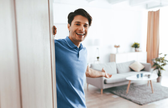 A cheerful man stands in the doorway of a well-lit living room. He smiles warmly, welcoming visitors into a space with modern furniture and plants, creating a friendly atmosphere. - Powered by Adobe