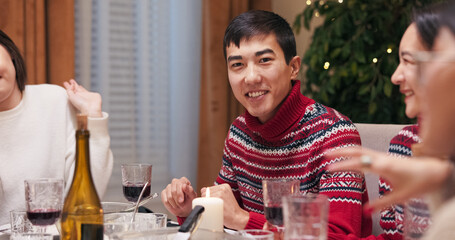 A young brunette man in a warm red sweater smiles at the camera from the festive table during a Christmas party. The guy looks at friends before posing, as the Christmas tree flickers behind him.