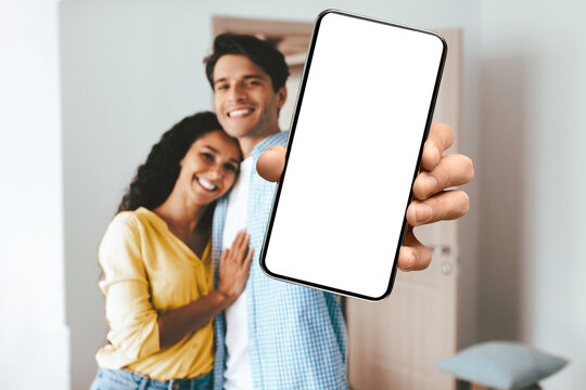 A joyful couple is smiling while embracing each other in a warm and inviting living room. The man holds up a smartphone in front of them with a blank screen. - Powered by Adobe