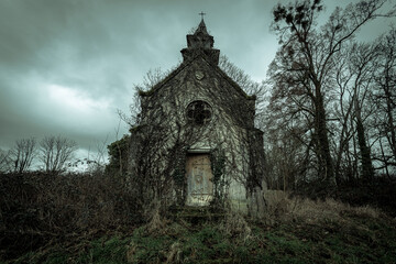 Vine-covered abandoned chapel with weathered wooden door and round window beneath a moody, overcast sky