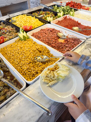 A child's hand scoops food onto a plate near a large table with corn, pasta and vegetable salads