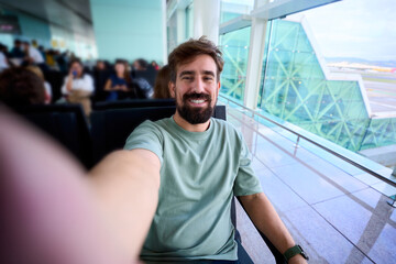Smiling caucasian millennial man taking a selfie while waiting at an airport gate, seated by large glass windows. Bright terminal architecture conveys travel mood and anticipation.