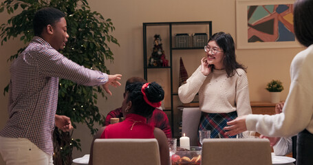 Guy in checkered shirt invited friends. They are sitting at the table, talking, smiling and having fun. The guy is standing and telling funny story and gesturing with hands.