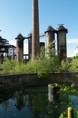 Abandoned industrial plant with rusty silos, tall chimney and overgrown vegetation reflected in a murky reservoir