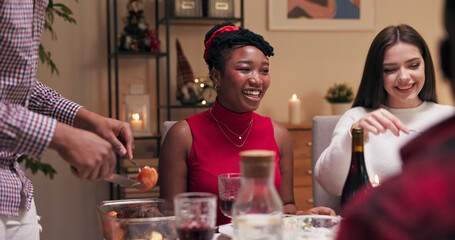 Sitting at the table and smiling during a friend's birthday celebration, are a girl with dark hair in a white sweater and a multi-ethnic girl in red clothes. A guy in a shirt cuts meat and puts it on