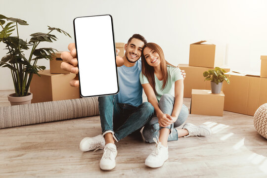 A cheerful couple sits on the floor of their new home surrounded by moving boxes. They are smiling and displaying a blank smartphone to capture the moment of joy and new beginnings together. - Powered by Adobe