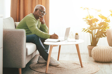 A senior man sits on a sofa, deep in thought while writing in a notebook. He uses a laptop on a coffee table, surrounded by plants and warm sunlight in a cozy room.