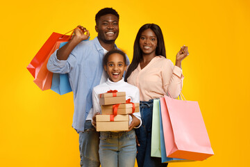 A joyful black family of three poses with colorful shopping bags and wrapped gift boxes. The happy parents and excited teen girl showcase their holiday spirit against a bright yellow background.