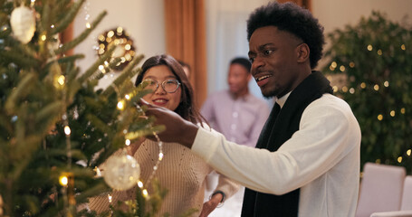 A conversation among friends takes place during a group dinner meeting. A woman with glasses is talking to a man with dark hair and stubble by a Christmas tree, telling each other about decorations