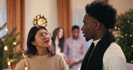Friends have a conversation during a team dinner meeting in a cozy living room. They stand by a Christmas tree adorned with baubles and lights, enjoying the decorated atmosphere.