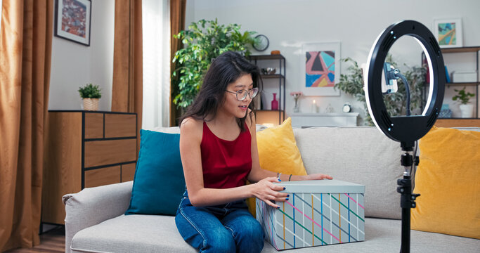 Happy young woman records unboxing content using her smartphone on a tripod. The beauty blogger unwraps cosmetics, describing textures and scents in her vlog. - Powered by Adobe