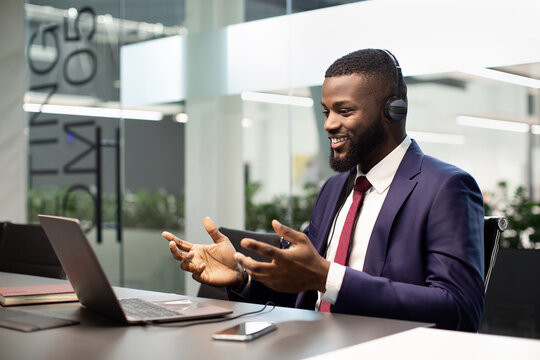 Happy young african american man CEO having video call at office, using headset and modern laptop, attending online conference or having video chat with business partners, copy space, side view