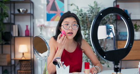 Beauty model applies foundation with a pink sponge while browsing in the mirror, recording makeup tips for her social media viewers.