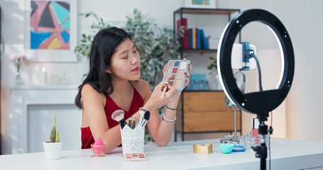 Experienced makeup artist of asian descent applies eye shadow on the eyelids, recording a tutorial on smoky eyes, in front of her on the table mirror, cosmetics and beauty accessories.