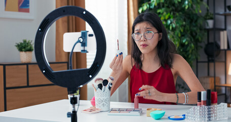 Happy beauty content creator working remotely, talking about matte lipsticks in front of smartphone. Woman opens product, shows details to viewers, filming at home.