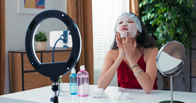 Woman applies face mask live on camera, explaining her skincare routine and makeup preferences. Ring light and smartphone support her beauty content creation.