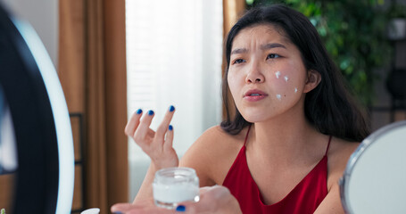 Woman with Korean features demonstrates facial skincare live on camera. She stands in front of a mirror, applies moisturizer and records beauty vlog at home.