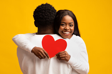 A joyful couple embraces, with the woman holding a red heart card behind her partner. They smile brightly, showcasing their affection in front of a vivid yellow backdrop.
