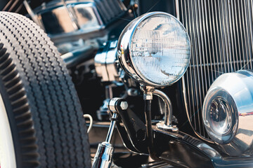 Headlight on vintage hot rod with vertical grille bars and classic tire visible. Front quarter view showing detailed automotive components including mounting bracket, bumper, and textured tire tread.