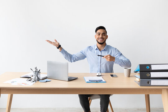 Cheerful bearded male is seated at a modern desk, smiling and pointing to an open area beside him. He holds a pen in one hand and looks inviting, ready to share ideas at his workspace.